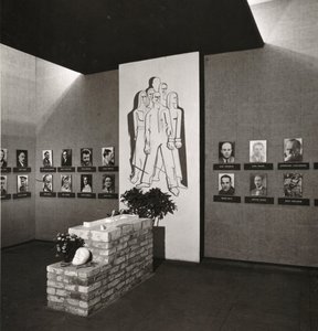 Blick in die Ausstellung: an einer Wand hängen historische Schwarz-Weiß-Porträts mit schwarzen Balken darunter. In der Mitte hängt ein weißes Tuch mit einem sechs abstrakt gemalten Menschen. Davor steht das Plötzensee-Modell.
